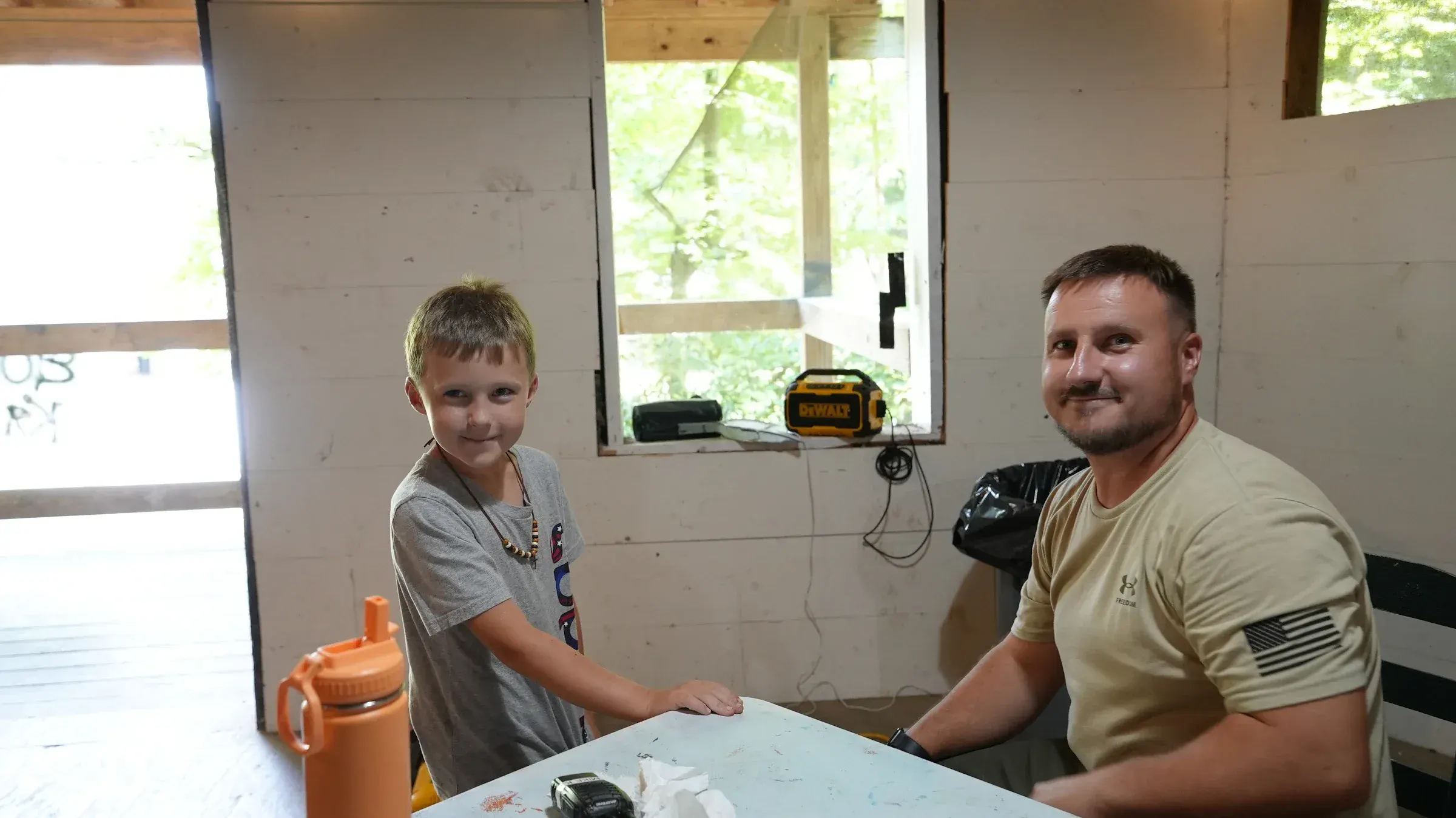 A young camper and his leader smile together at a cabin table.